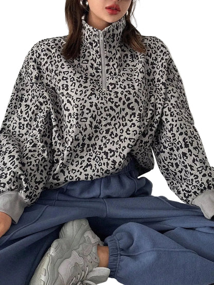 A person wearing a black and white leopard print half-zip pullover sweater, paired with blue sweatpants and white sneakers, sitting against a plain light background.