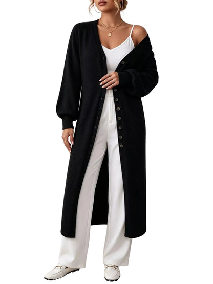 A woman models a long black button-front cardigan over a white camisole and white wide-leg pants, standing against a textured white wall on a white shag rug.