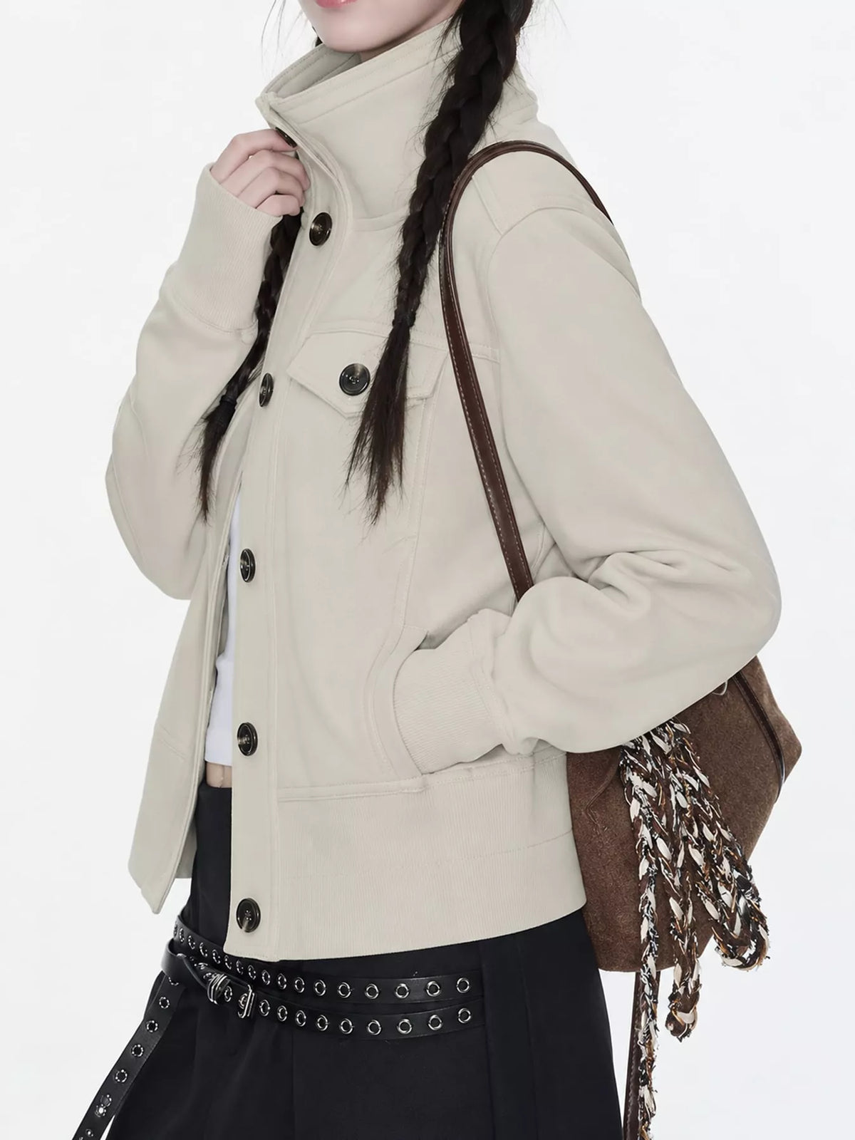 A model wearing a beige high-collar utility jacket with dark buttons, styled with a brown shoulder bag and a black skirt with silver studs.