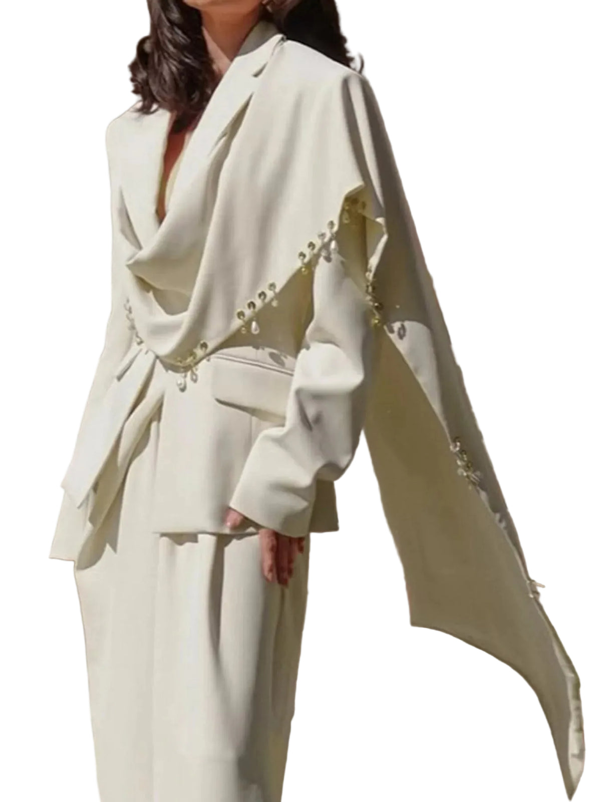 A model wearing a cream-colored tailored suit with a draped cape-style jacket and matching trousers, featuring decorative bead details along the sleeve edge, set against a beige stone wall background.