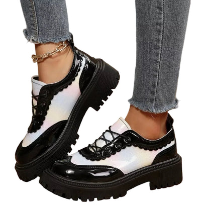 A pair of chunky low-top lace-up shoes with glossy black patent leather panels, white scalloped overlays, and thick black lug soles, shown worn with gray frayed-hem jeans on a rustic wooden floor.