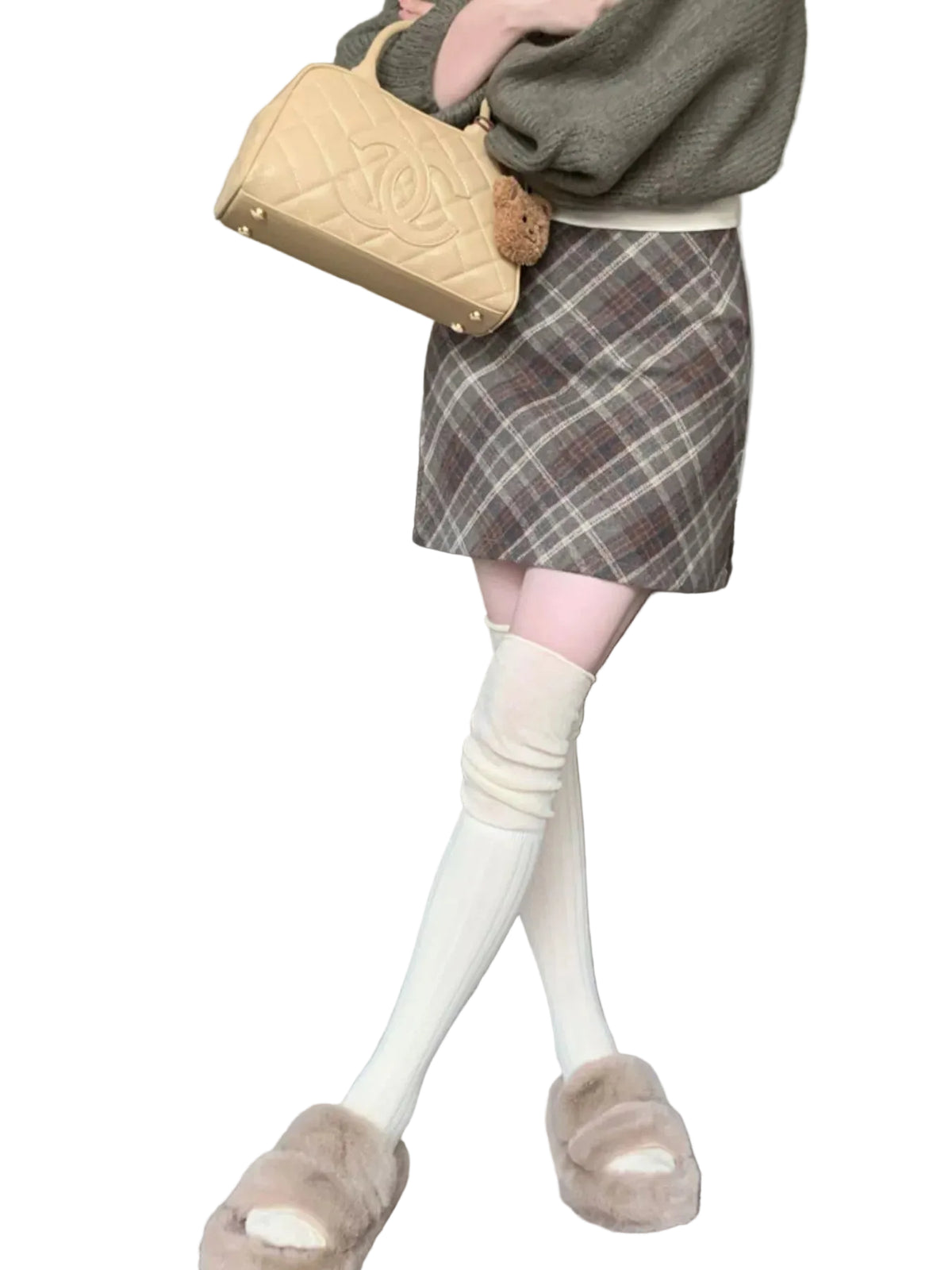 A person standing indoors wearing a brown plaid mini skirt, cream knee-high socks, beige slippers, and holding a beige quilted handbag. A gray sofa and carpeted room are visible in the background.