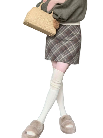 A person standing indoors wearing a brown plaid mini skirt, cream knee-high socks, beige slippers, and holding a beige quilted handbag. A gray sofa and carpeted room are visible in the background.