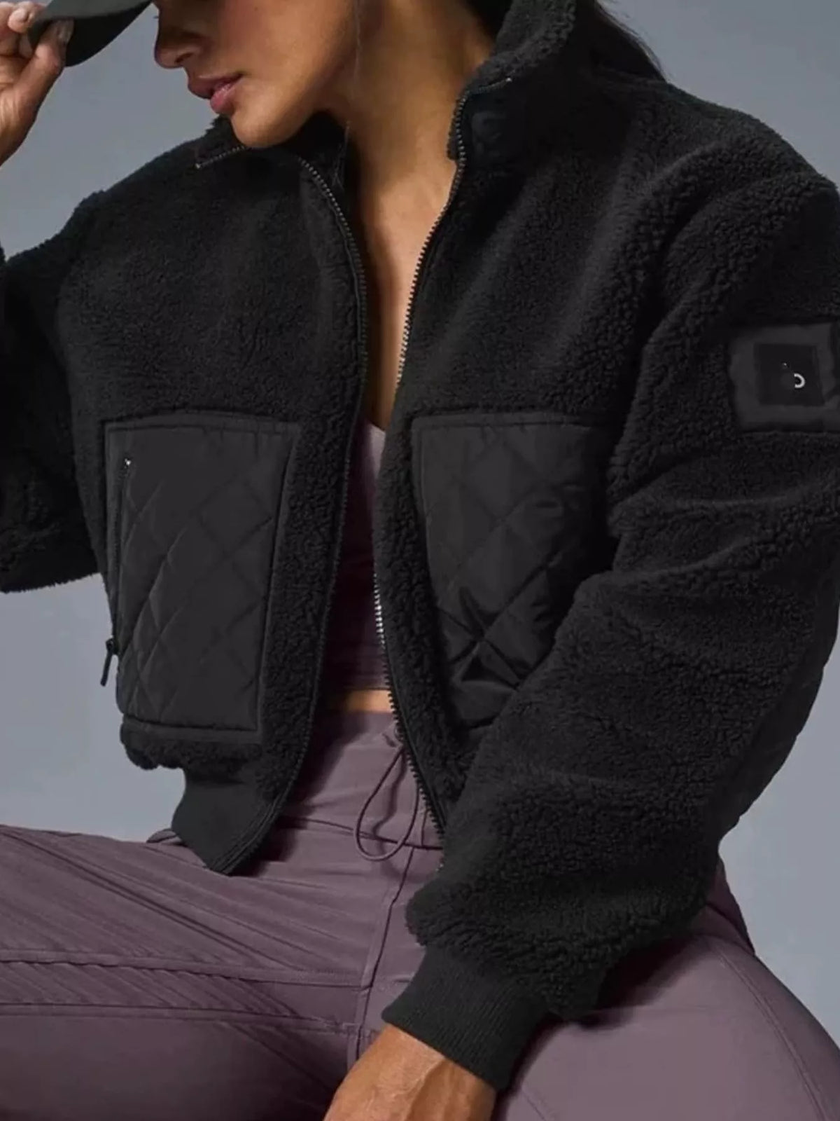 A close-up of a woman wearing a black fleece zip-up jacket with quilted front pockets and a small logo patch on the sleeve, paired with mauve athletic pants against a gray studio background.