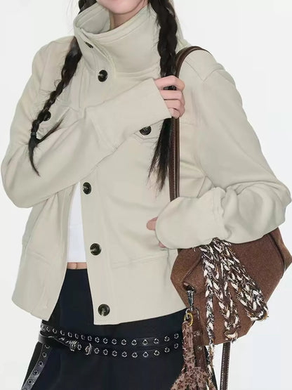 A model wearing a cream-colored high-neck utility jacket with black buttons, styled with a brown shoulder bag and a studded belt.