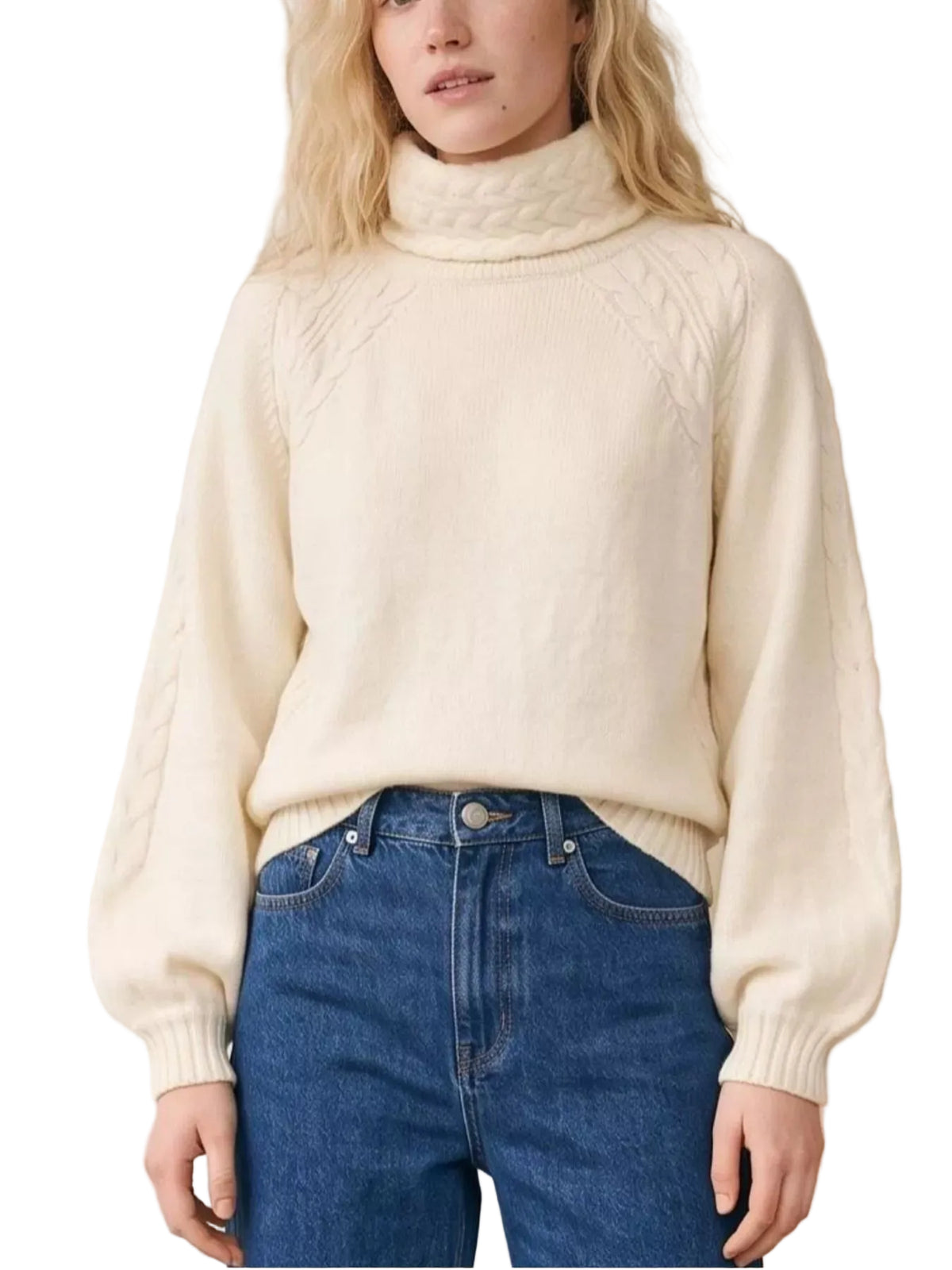 A woman wearing a cream-colored cable-knit mock neck sweater and blue jeans, standing in front of a white brick wall.