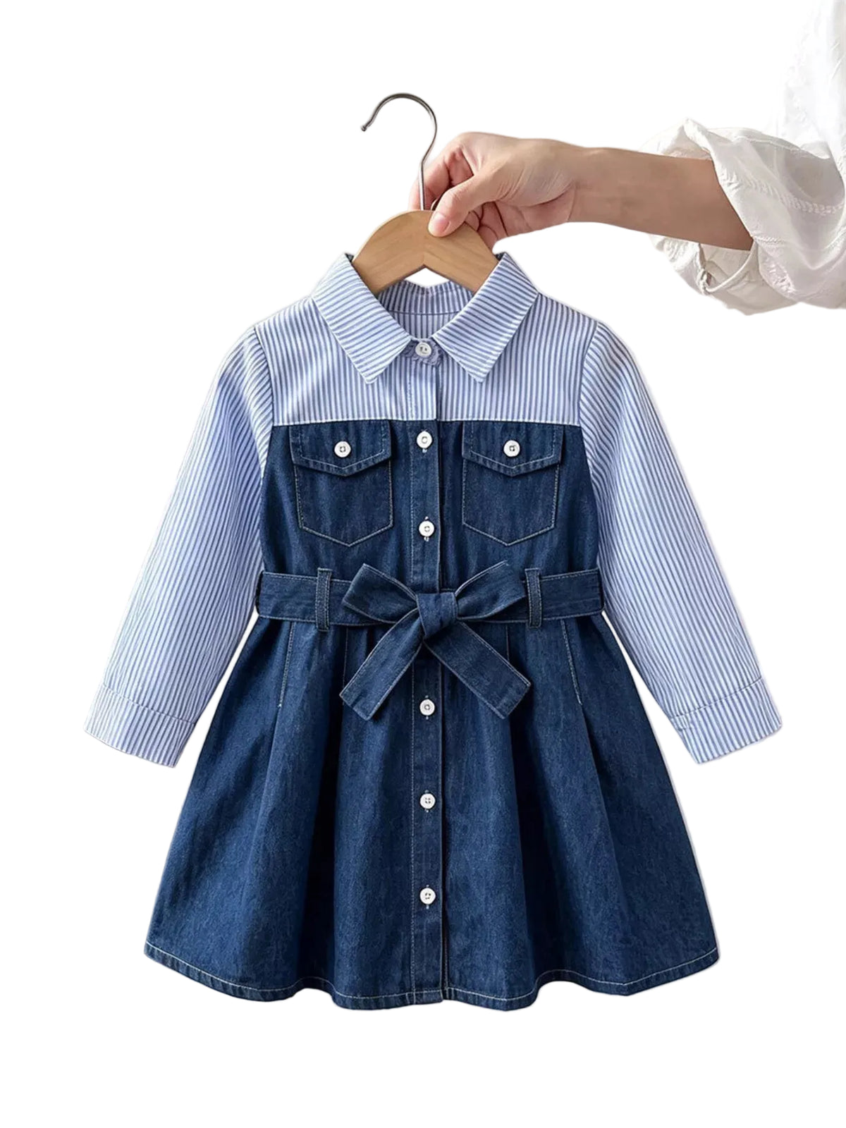 A toddler girl's dress hanging on a wooden hanger, held by a hand. The dress features a light blue and white striped long-sleeve shirt collar and upper body, and a dark blue denim lower body with a matching denim tie belt.