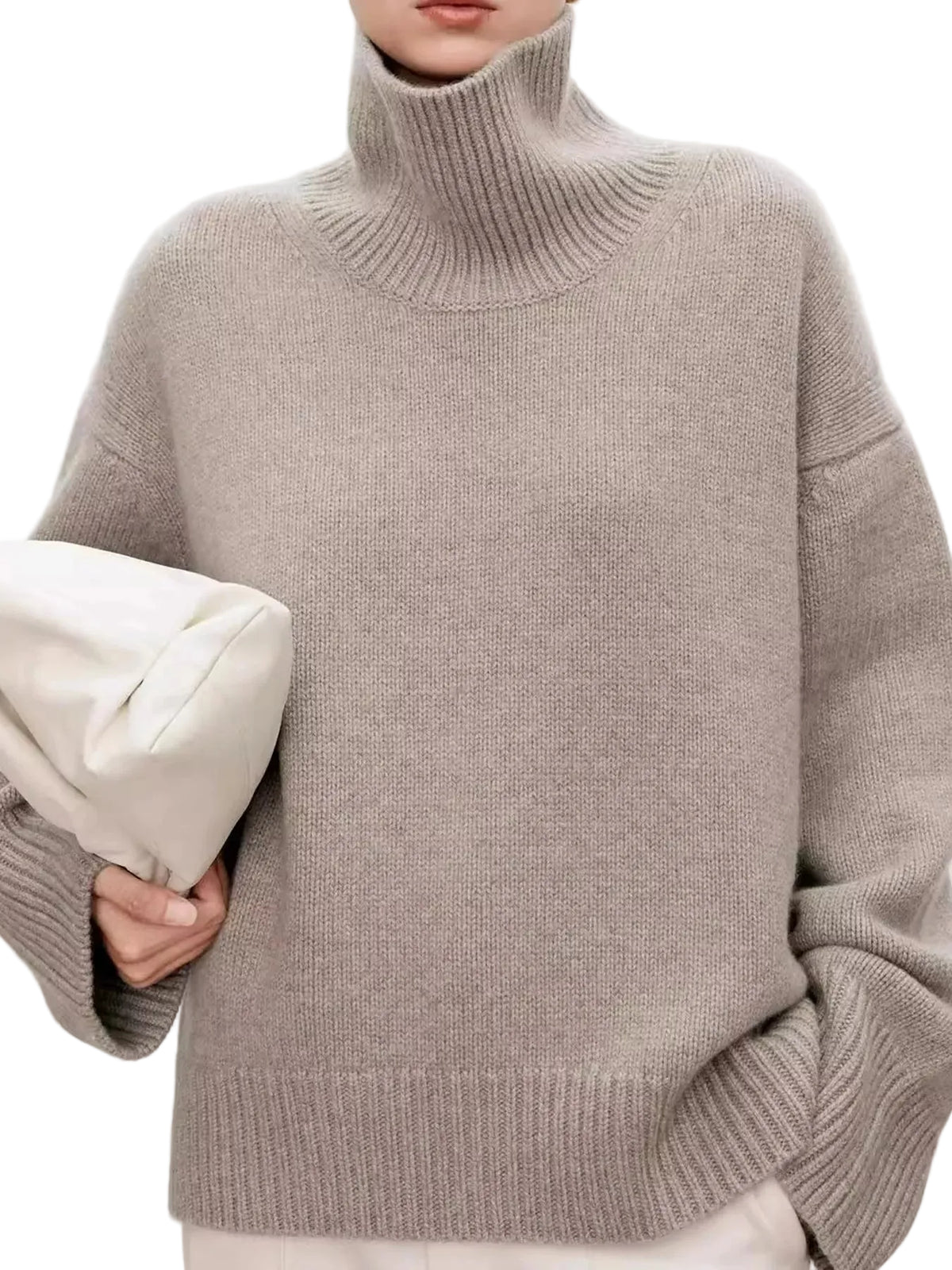 A person wearing an oversized ribbed turtleneck sweater in a light taupe-beige color, holding a white envelope-style clutch against a plain light background.