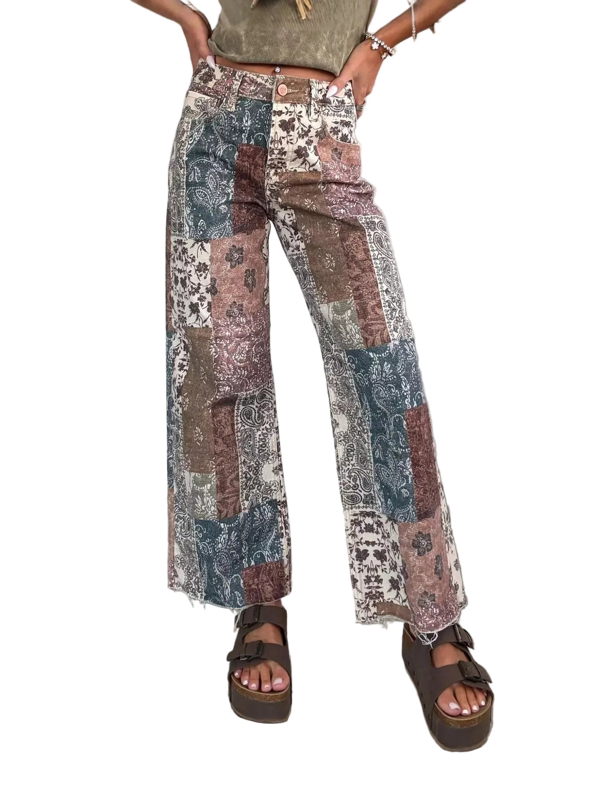 A person wearing patchwork wide-leg pants with a distressed bohemian pattern in muted brown, blue, beige, and cream tones, paired with brown platform sandals against a textured gray wall and tiled floor.