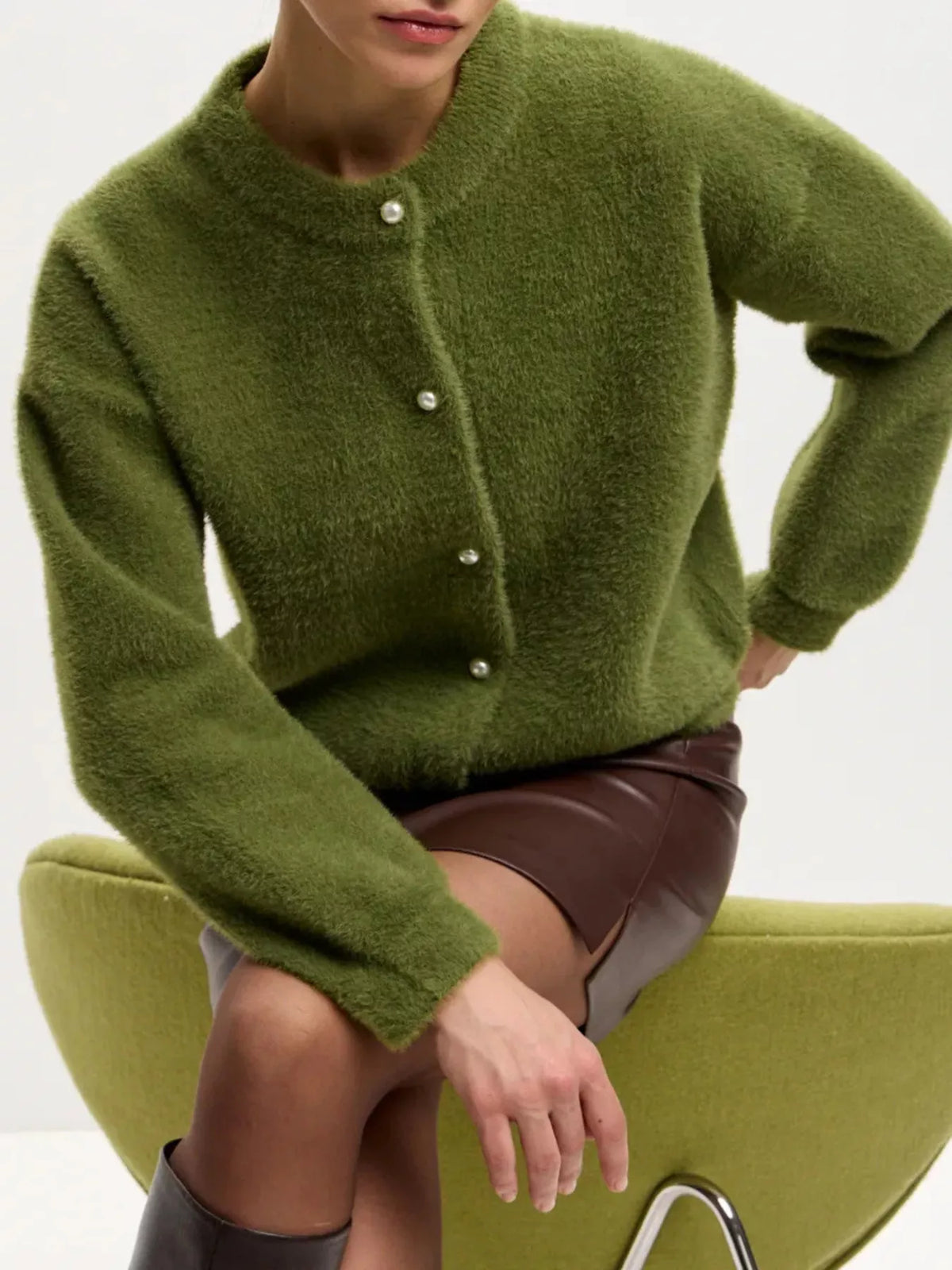 A model wearing a fuzzy olive green button-up cardigan sweater, seated on a matching green chair against a plain white background.