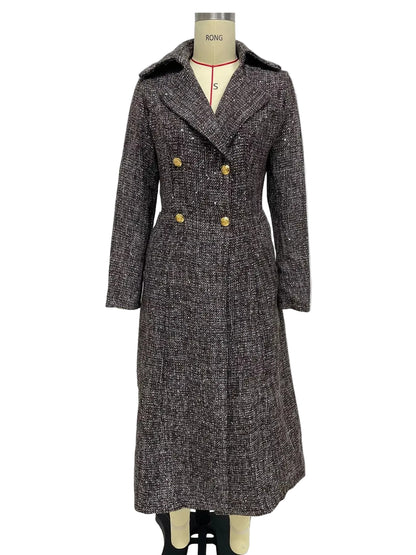 A long double-breasted tweed coat displayed on a mannequin against a plain light gray wall. The coat has a dark gray and black textured tweed pattern with prominent gold buttons and a wide collar.