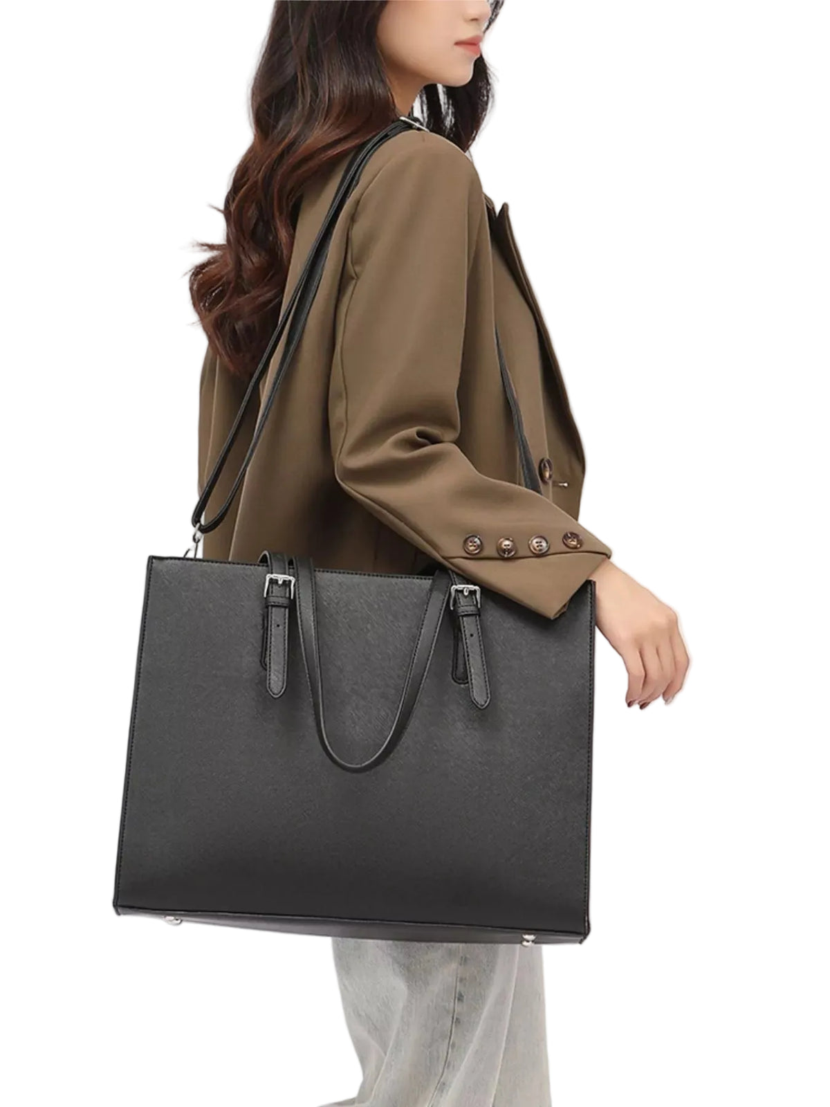 A woman wearing a brown blazer and light gray jeans carries a large black tote bag with a shoulder strap and top handles against a plain white background.
