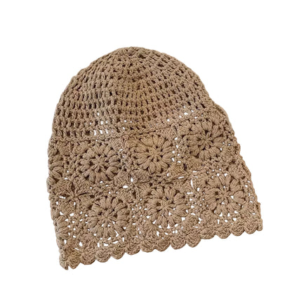 A beige crocheted beanie hat with a scalloped edge and floral granny-square pattern, laid flat on a wooden surface with a cream fabric and woven accessory in the background.