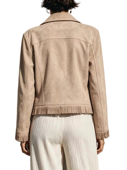A person shown from the back wearing a light beige suede jacket with fringe trim at the hem and cuffs, paired with white pleated trousers, standing against a textured wall with a strong shadow.