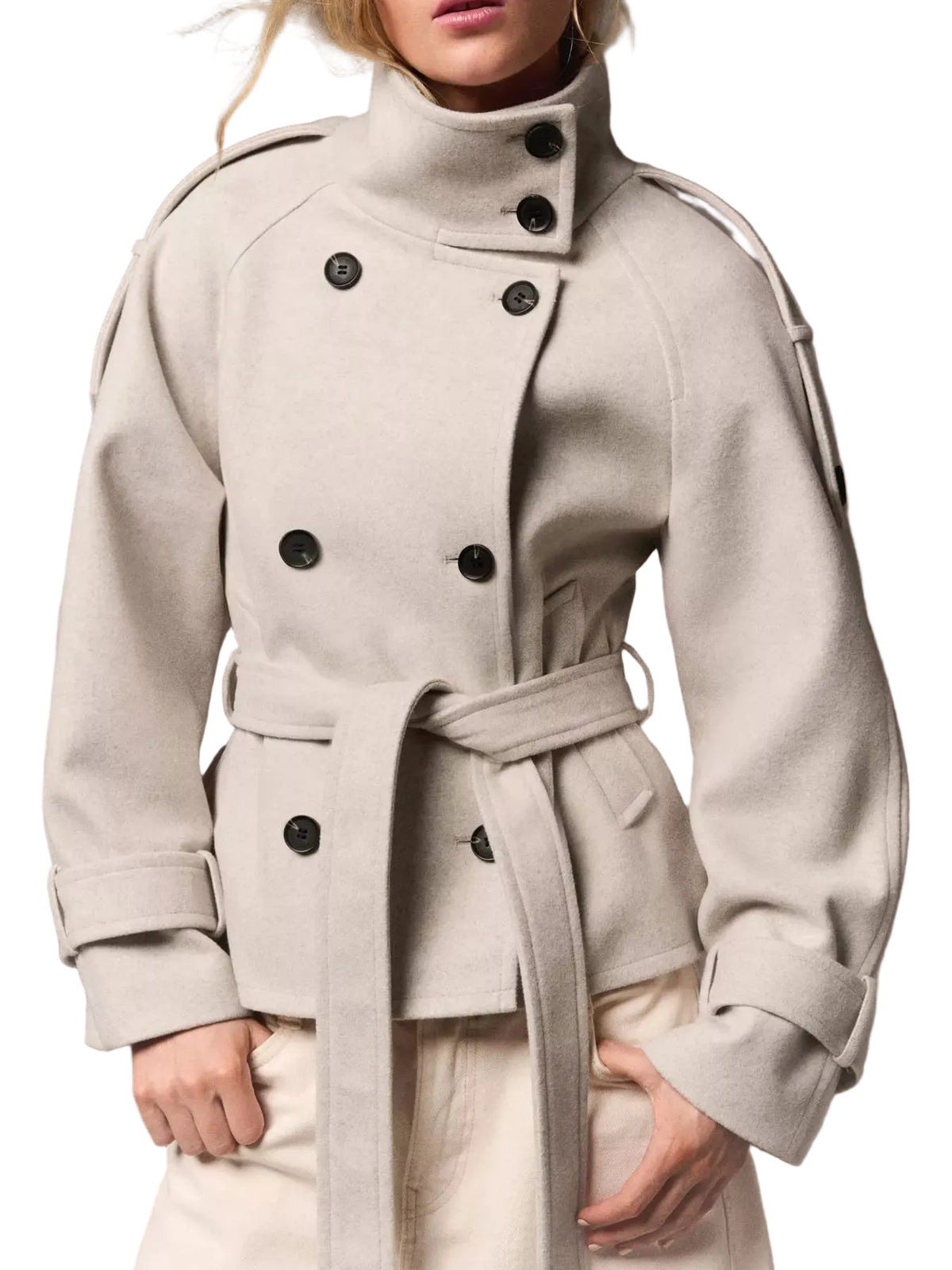 A model wearing a light beige double-breasted belted coat with black buttons, paired with cream trousers, against a neutral studio background.