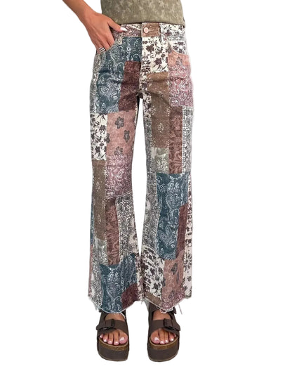A person wearing patchwork wide-leg pants with a mix of blue, brown, beige, and white patterns, paired with brown platform sandals against a textured gray wall and tiled floor.