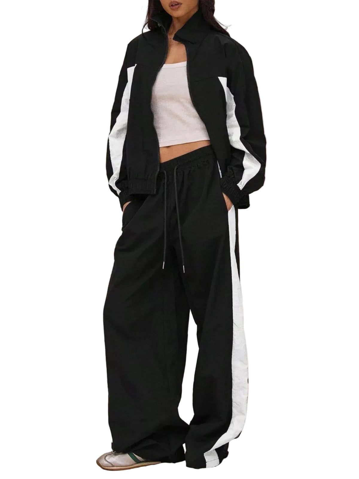A model wearing a black and white athletic tracksuit consisting of a zip-up jacket and matching wide-leg sweatpants, styled with a white crop top and sneakers against a solid beige background.