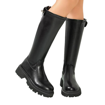 A pair of black knee-high leather boots with chunky lug soles, shown on a model's legs in a bright, modern indoor setting.