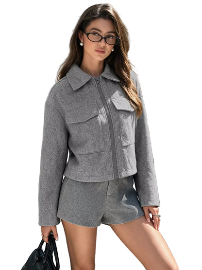 A woman wearing a gray cropped zip-front jacket and matching gray shorts, standing outdoors on a city sidewalk in front of a stone building.