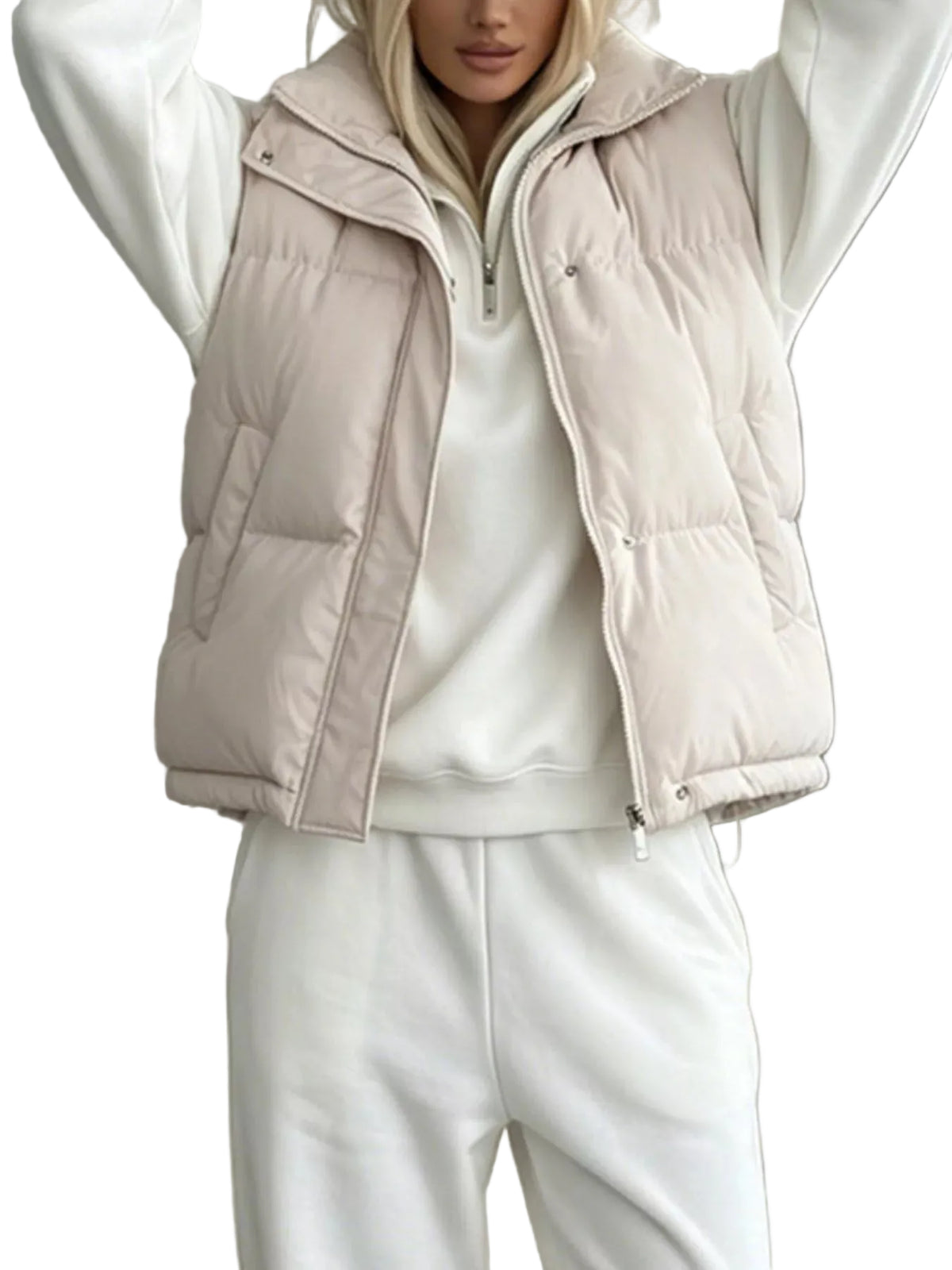 A woman wearing a light beige puffer vest over a white zip-up sweatshirt and white sweatpants, with her arms raised in front of a neutral gray wall and white sofa.