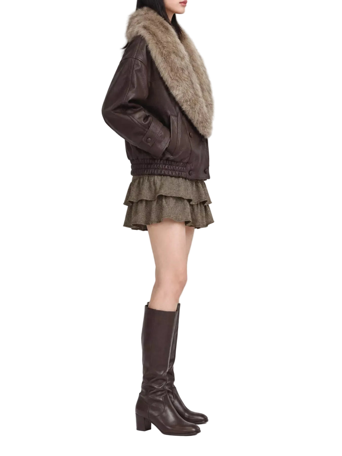 A model shown from the side wearing a dark brown leather jacket with a large beige fur collar, a brown ruffled mini skirt, and dark brown knee-high leather boots, set against a plain light gray studio background.