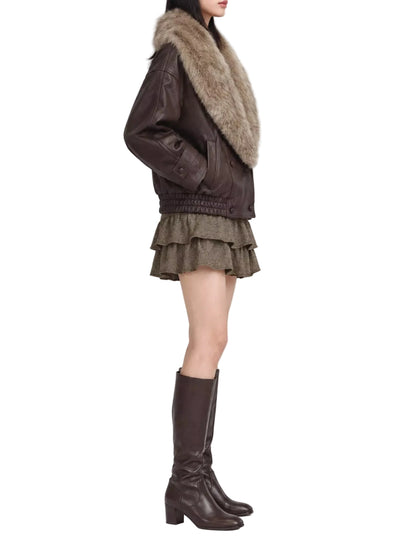 A model shown from the side wearing a dark brown leather jacket with a large beige fur collar, a brown ruffled mini skirt, and dark brown knee-high leather boots, set against a plain light gray studio background.