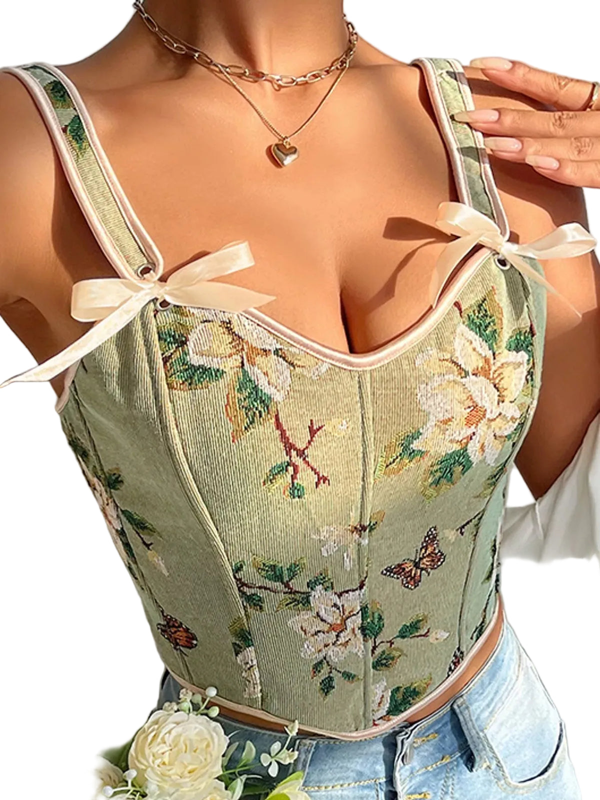 A close-up of a woman wearing a light sage green floral corset-style camisole top with cream ribbon bows at the straps, paired with light blue denim shorts.