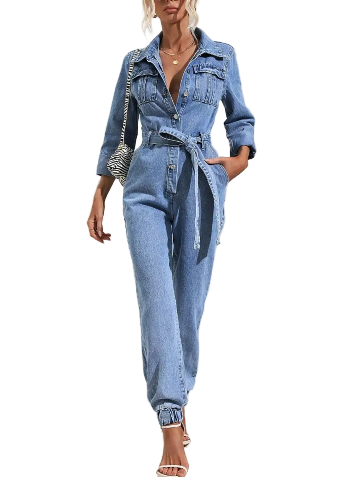 A woman models a light blue denim one-piece jumpsuit with a button-up front, chest pockets, rolled sleeves, a matching waist tie belt, and cuffed ankles, styled with a patterned shoulder bag and white high-heeled sandals against a white wall with palm leaf shadows.