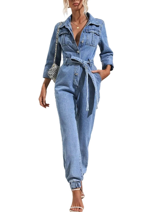 A woman models a light blue denim one-piece jumpsuit with a button-up front, chest pockets, rolled sleeves, a matching waist tie belt, and cuffed ankles, styled with a patterned shoulder bag and white high-heeled sandals against a white wall with palm leaf shadows.