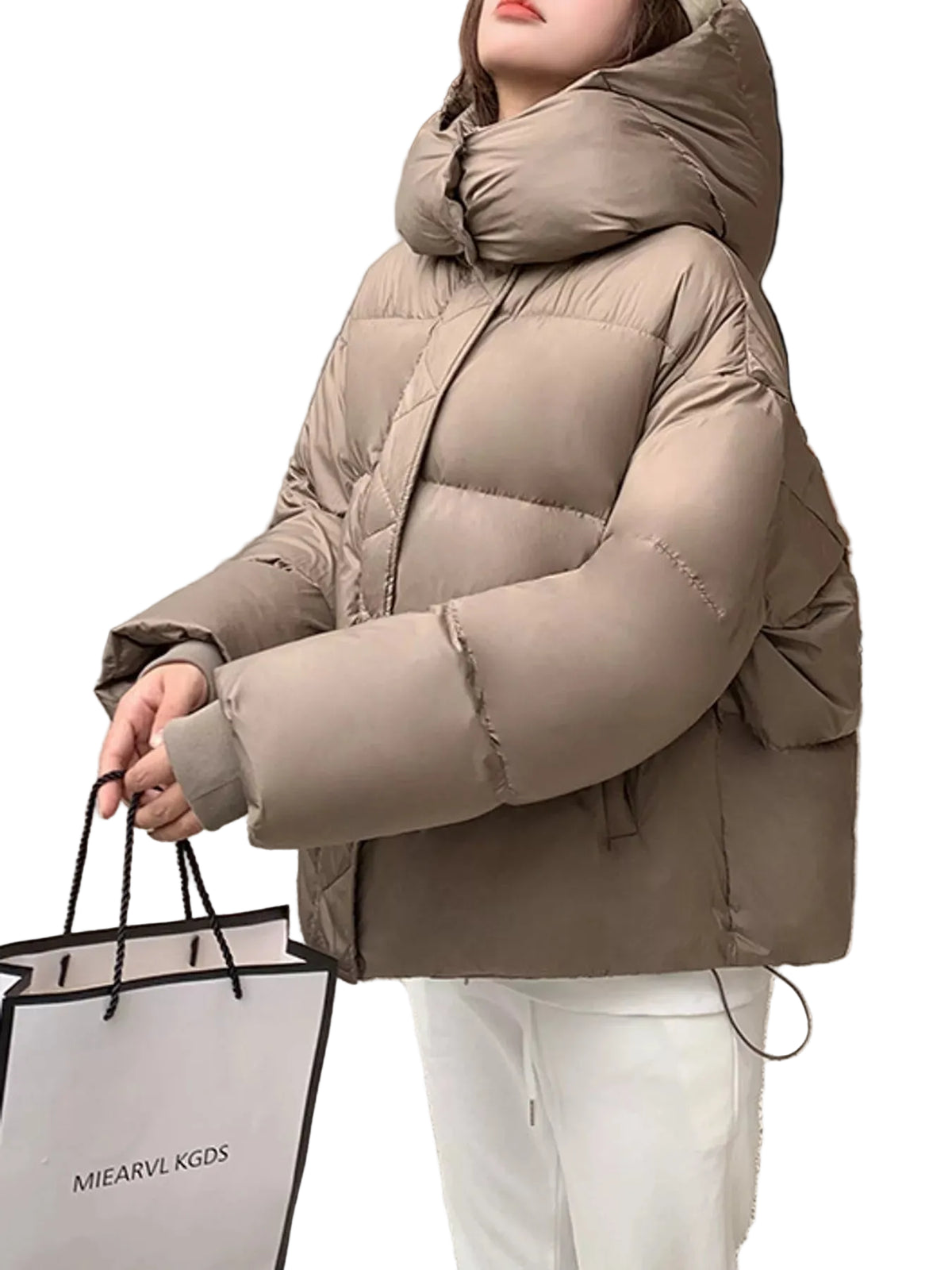 A woman wearing a beige oversized puffer jacket with a high padded collar, holding a white shopping bag with the brand name MIEARVL KGDS.