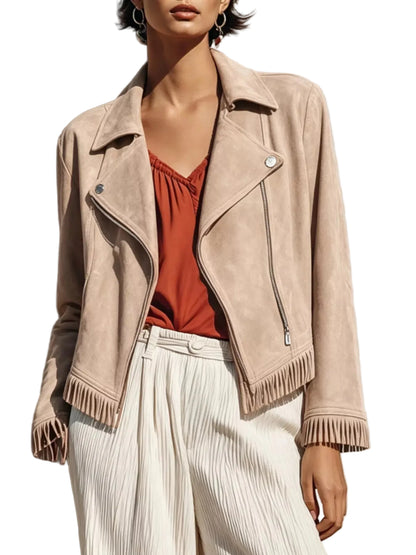A woman models a beige suede moto jacket with fringe details, worn over a rust-orange top and white pleated trousers, set against a textured concrete wall with strong sunlight and shadows.