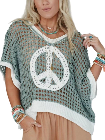 A woman wearing a loose, open-knit crocheted poncho top in sage green and white, featuring a large white peace sign motif on the front. She is also wearing layered turquoise necklaces and colorful beaded bracelets, paired with tan pants, against a plain white background.
