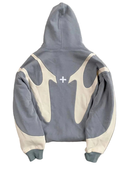 A gray and cream color-blocked hoodie laid flat on a textured gray concrete surface, showing the back view with a small white cross graphic in the center.