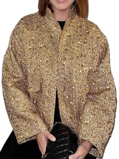 A woman wearing a cropped, oversized gold sequined jacket with a stand collar and front opening, paired with a black top and black quilted handbag against a plain white background.