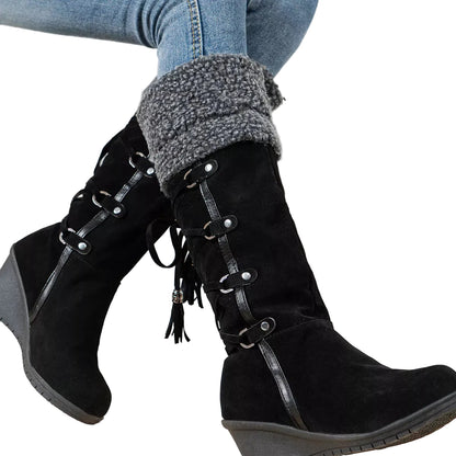 A pair of black mid-calf wedge boots with faux-leather lace-up detailing, tassel accents, and a gray fuzzy cuff, shown on a person wearing blue jeans.