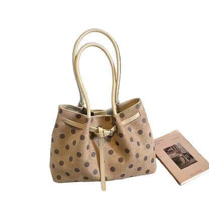 A beige suede-like tote bag with dark brown polka dots and light beige handles, placed on a textured gray carpet next to a book, with a black bench in the background.
