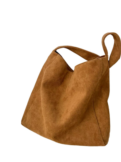 A large slouchy suede tote bag in a warm camel brown color, resting on a white surface next to a small ceramic cup and saucer against a white wall.
