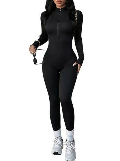 A woman modeling a black long-sleeve zip-up bodysuit with matching leggings, styled with white socks and white sneakers, standing outdoors next to a white wall.