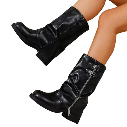 A pair of black leather mid-calf boots with side zippers and buckle details, shown on a model's legs against a textured gray wall and concrete surface.