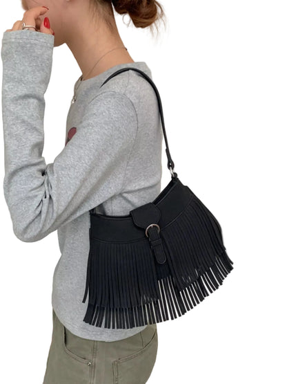 A woman wearing a gray long-sleeve top and olive green pants carries a black fringe shoulder bag with a wide black belt detail and silver-tone buckle.