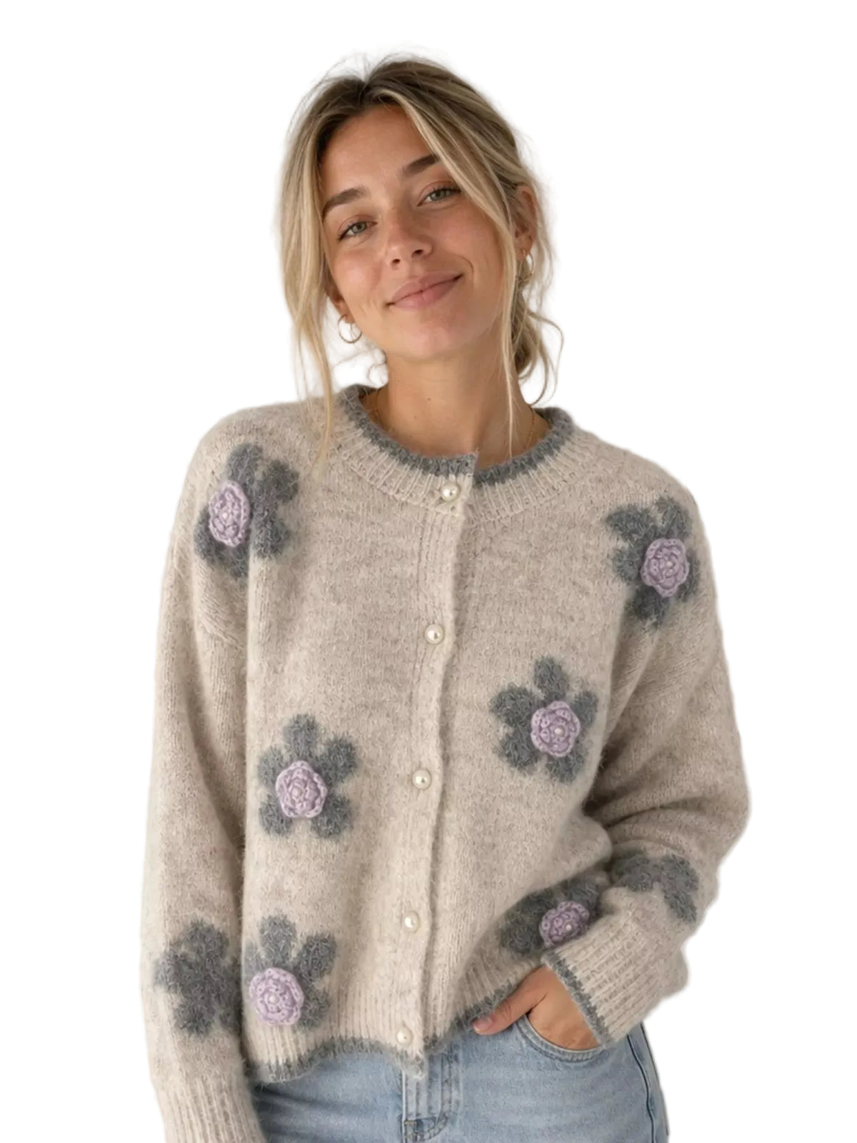 A woman wearing a beige fuzzy knit cardigan with large gray and lavender crocheted floral motifs, paired with light blue jeans in a bright indoor setting.
