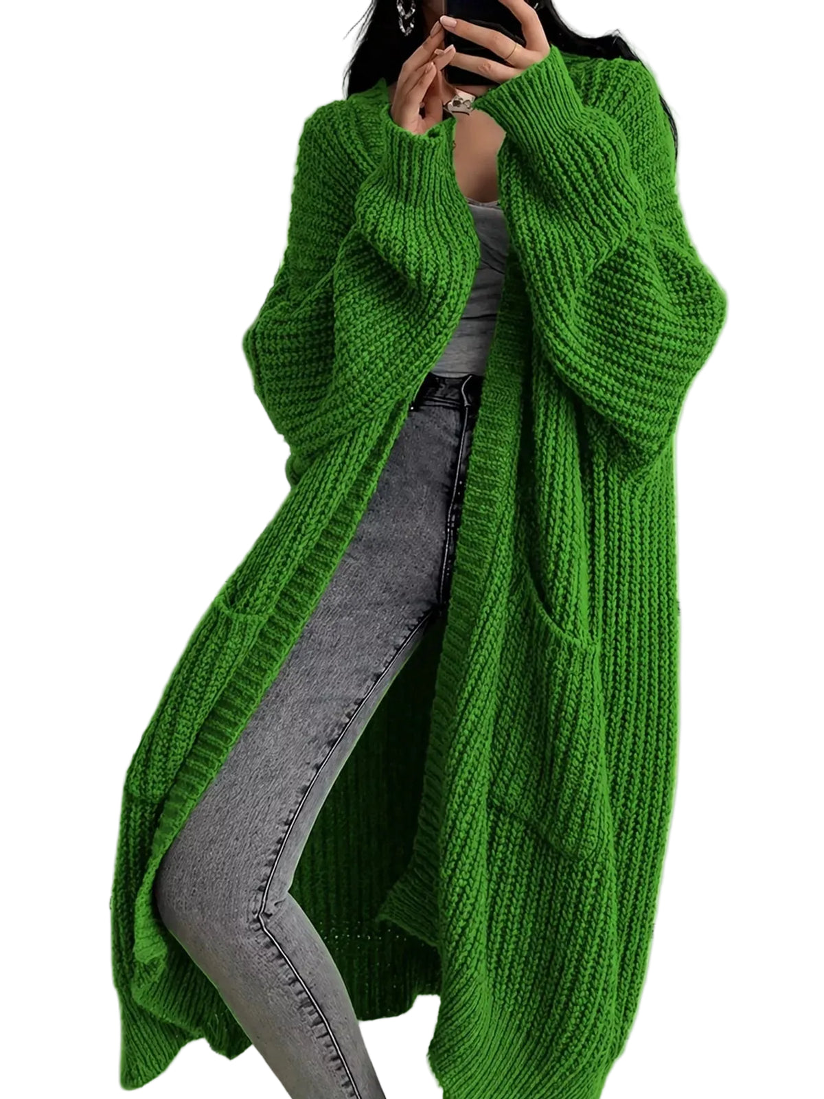 A person wearing an oversized bright green chunky knit cardigan over a gray top and gray jeans, standing indoors near a white shelf with a beige tote bag.