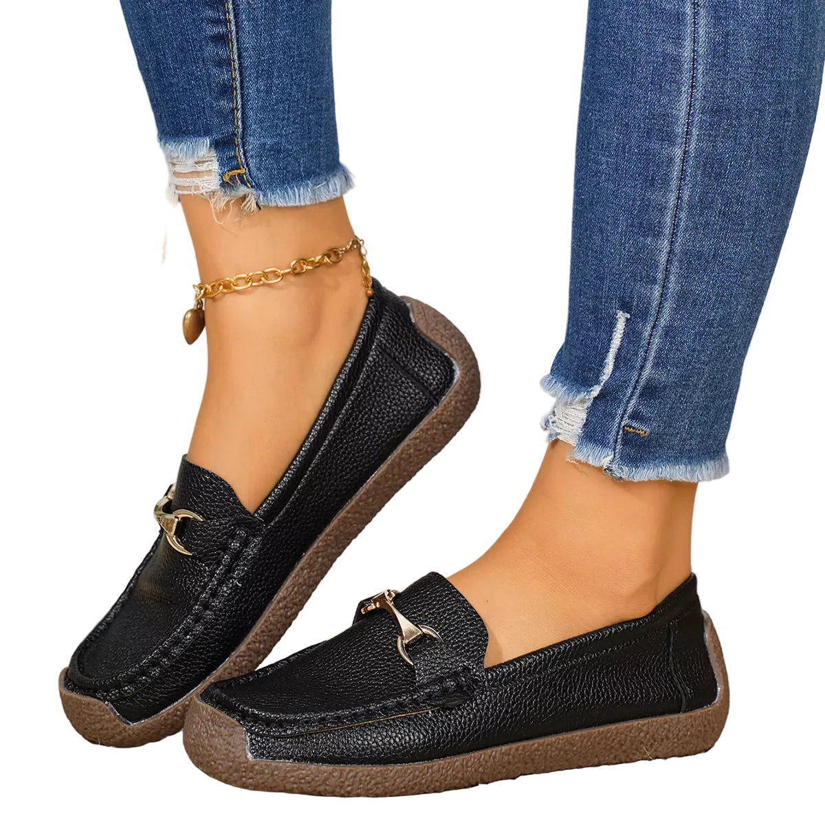 A person wearing black pebbled leather slip-on loafers with gold-tone horsebit details, paired with frayed blue jeans and a gold anklet, standing on a woven straw mat.