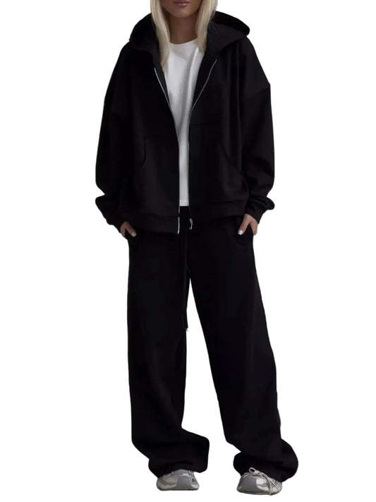 A model wearing a black zip-up hoodie, black wide-leg sweatpants, and a white t-shirt, standing in front of a light gray sofa.