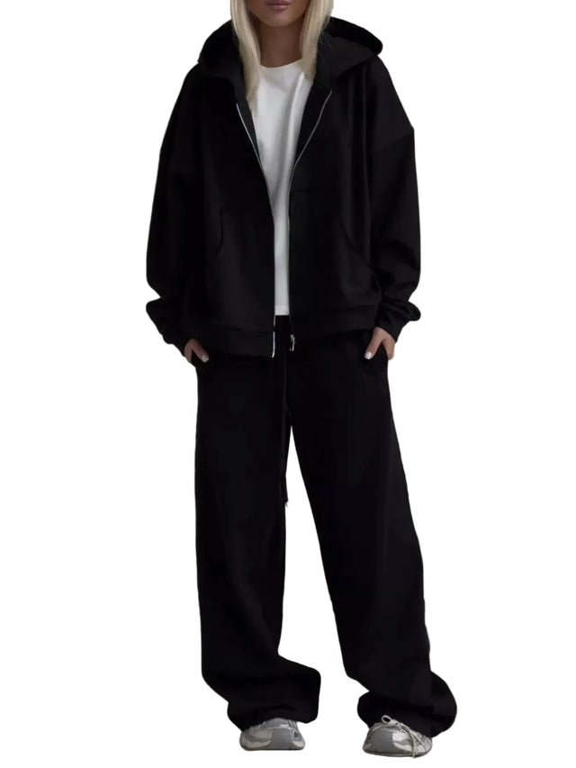 A model wearing a black zip-up hoodie, black wide-leg sweatpants, and a white t-shirt, standing in front of a light gray sofa.