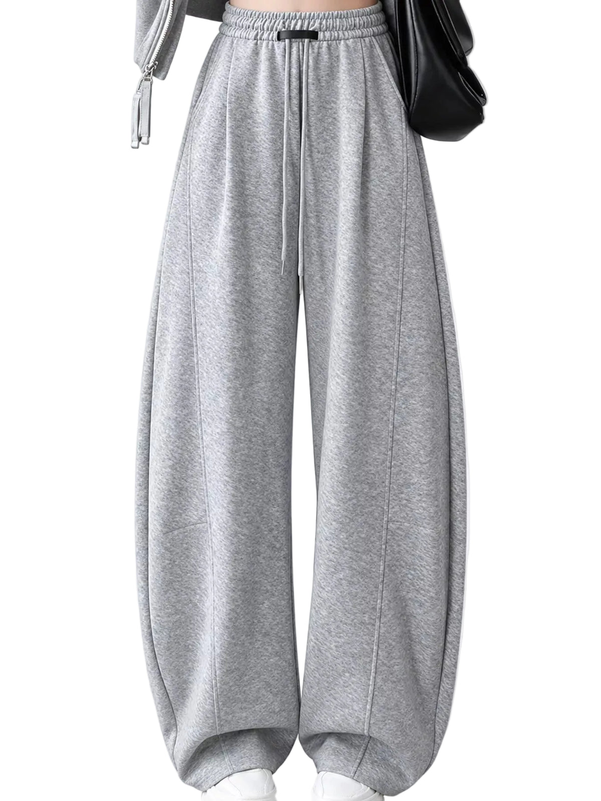 A person wearing loose, wide-leg gray sweatpants with an elastic waistband and drawstring, shown from the torso down against a light gray studio background. A black shoulder bag and white shoes are also visible.