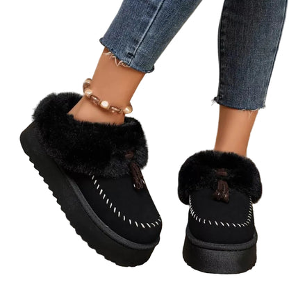 A person wearing black platform slip-on slippers with fluffy black faux-fur trim and white contrast stitching, paired with blue jeans and a pearl anklet, standing on a woven rug.