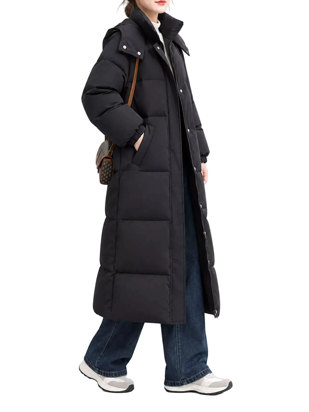 A model wearing a long black puffer coat over a white top, blue jeans, and white sneakers, carrying a small brown patterned shoulder bag against a light gray studio background.