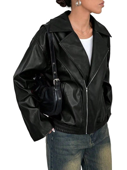 A woman wearing a black faux-leather moto jacket with silver zippers, styled with a white top, blue jeans, and a black shoulder bag against a light gray background.