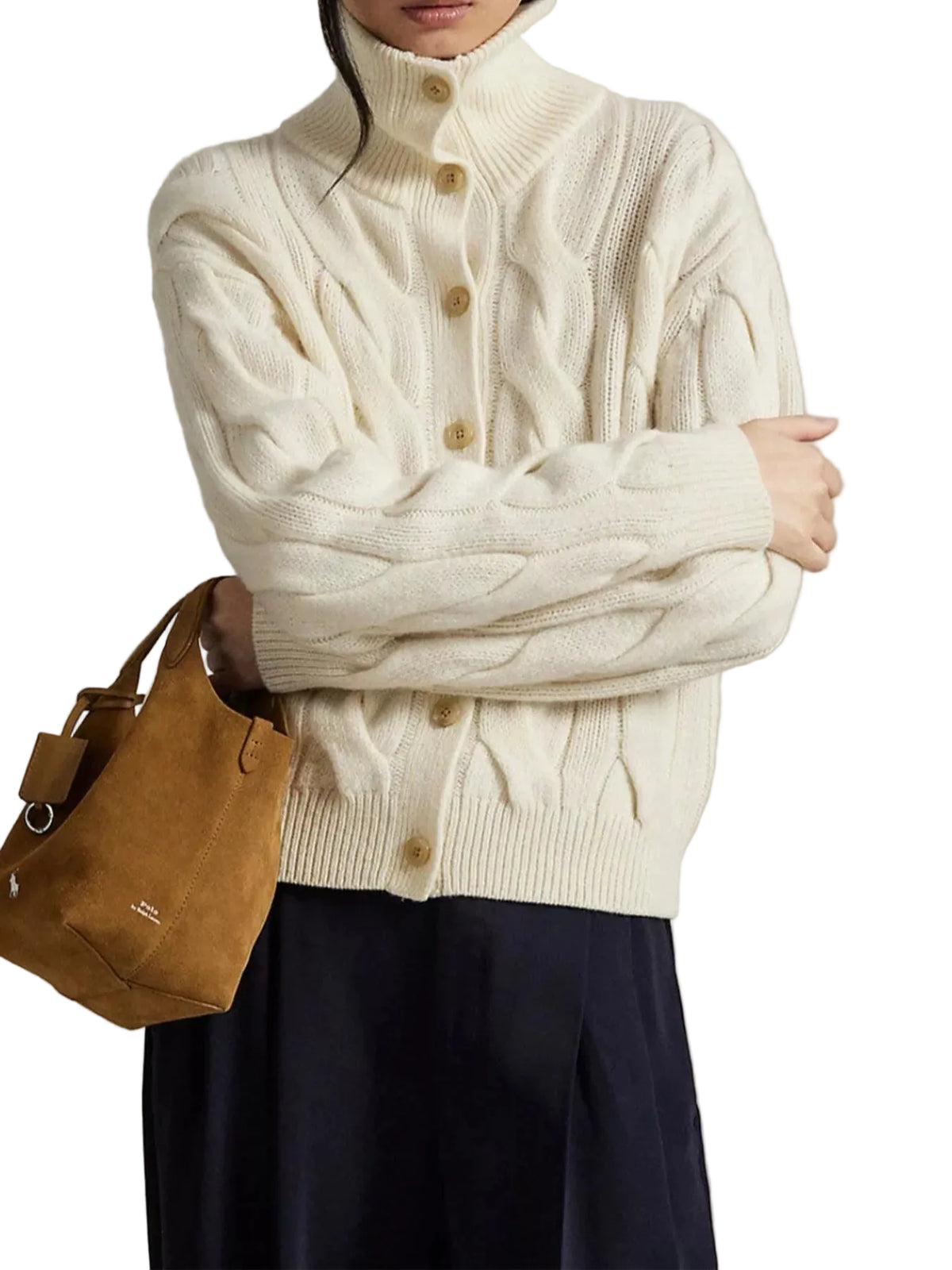A person wearing a cream cable-knit turtleneck cardigan with brown buttons, paired with dark navy pants and holding a tan suede shoulder bag against a neutral beige background.